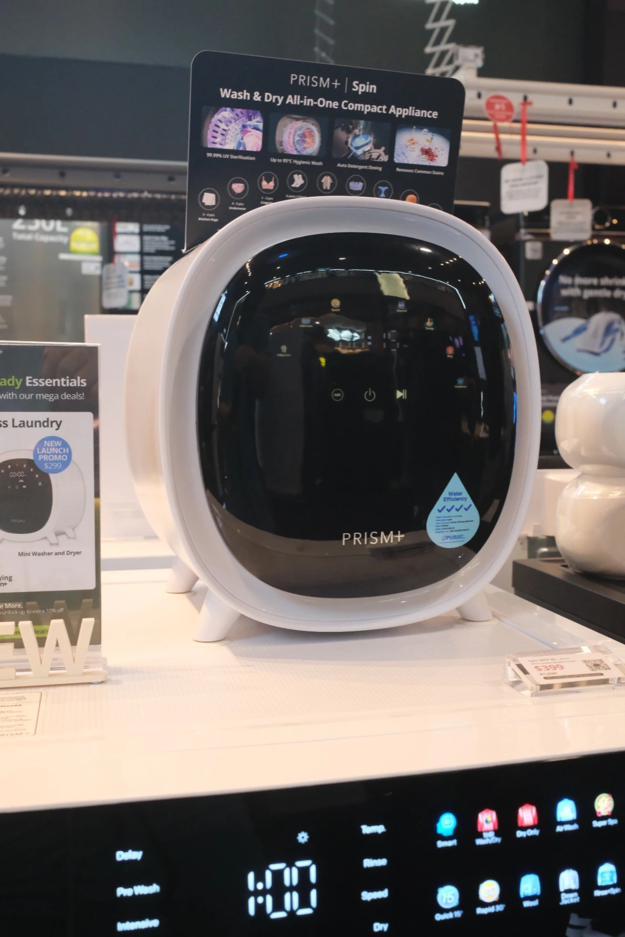 White PRISM+ Spin Mini washer-dryer on a retail display above a full-size washing machine control panel.